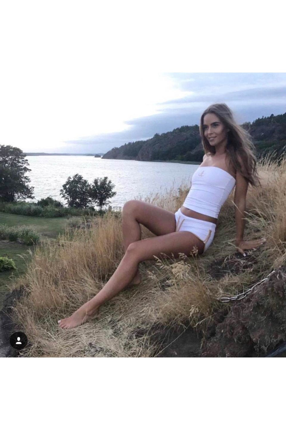 Abendlicht am Wasser. Das Model sitzt im hohen Gras im weißen Bandeau-Top und weißen Shorts.