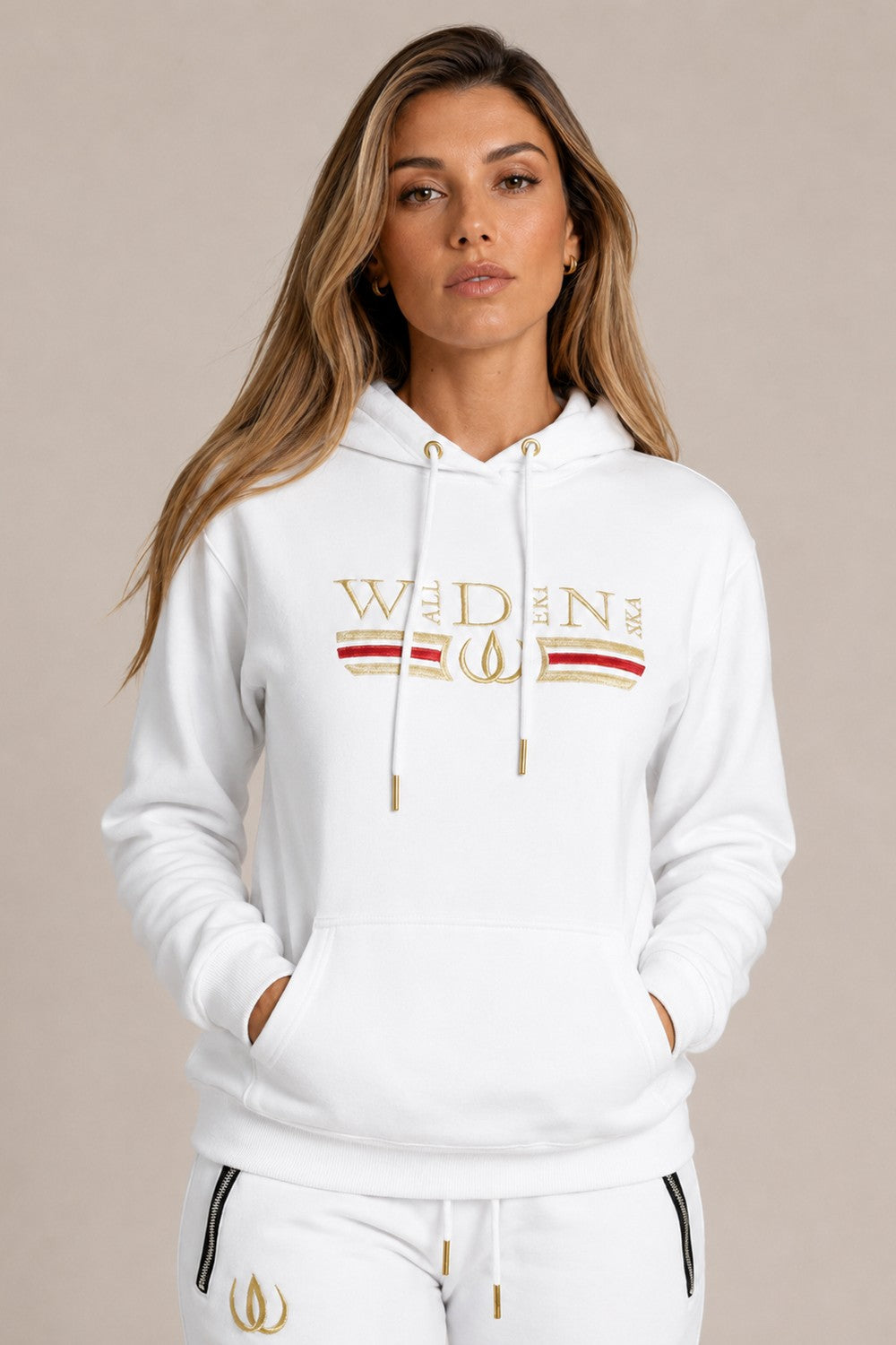 Weißer Deluxe Hoodie mit Goldstickerei und rotem Streifen auf der Brust, getragen zu weißen Deluxe Joggers mit Goldlogo.