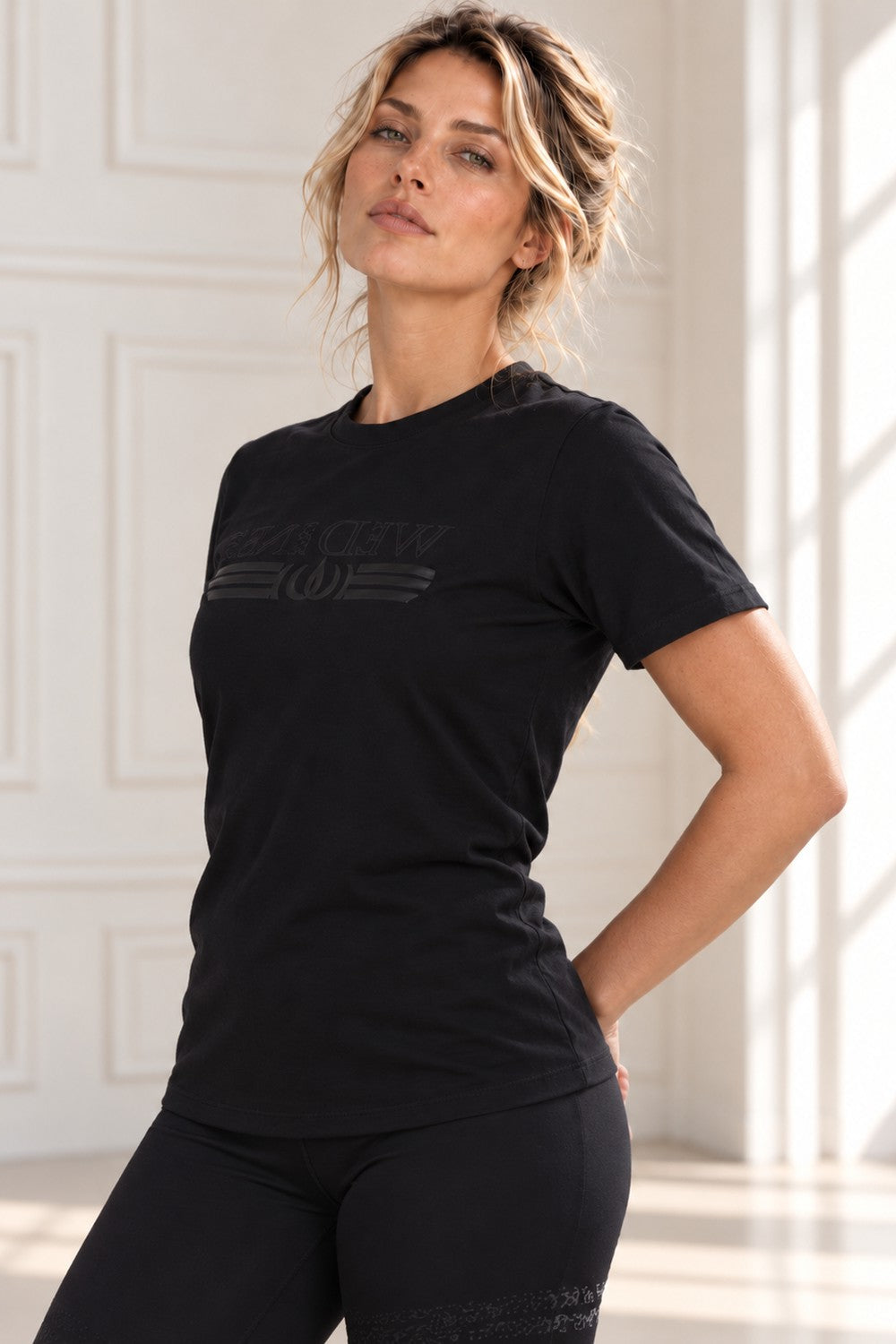 Das Model steht mit der Hand in die Hüfte gestützt im schwarzen Top mit Ton-in-Ton-Logo.