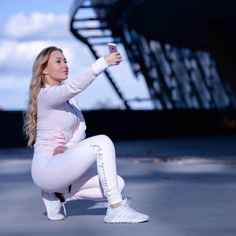 Draußen auf einem Dach sitzt das Model in der Hocke in der hellrosa Trainingshose und passenden Jacke.
