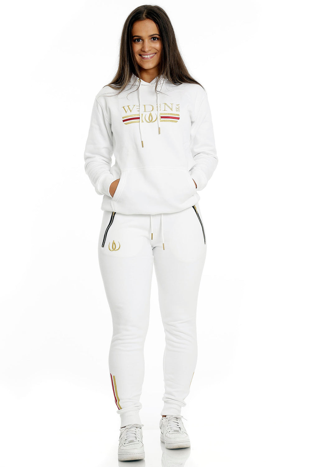 Stehend in weißen Joggern mit Goldlogo und weißem Hoodie mit Goldstickerei, zu weißen Sneakern.