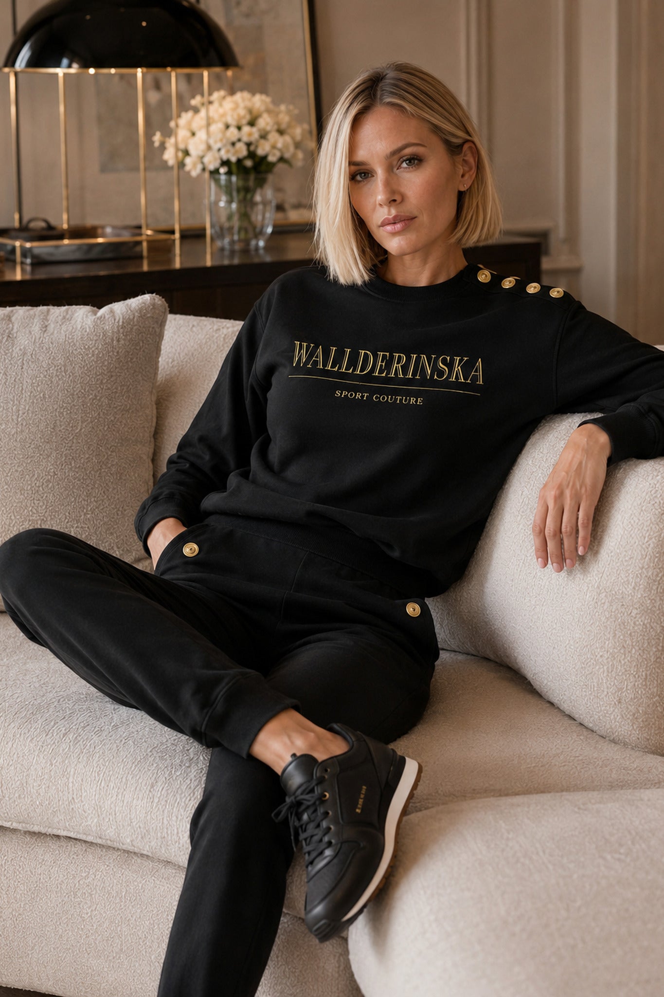 Edge Gold Button Sweater i svart med guldtext på bröstet och guldknappar på axeln, buren i en ljus soffa.