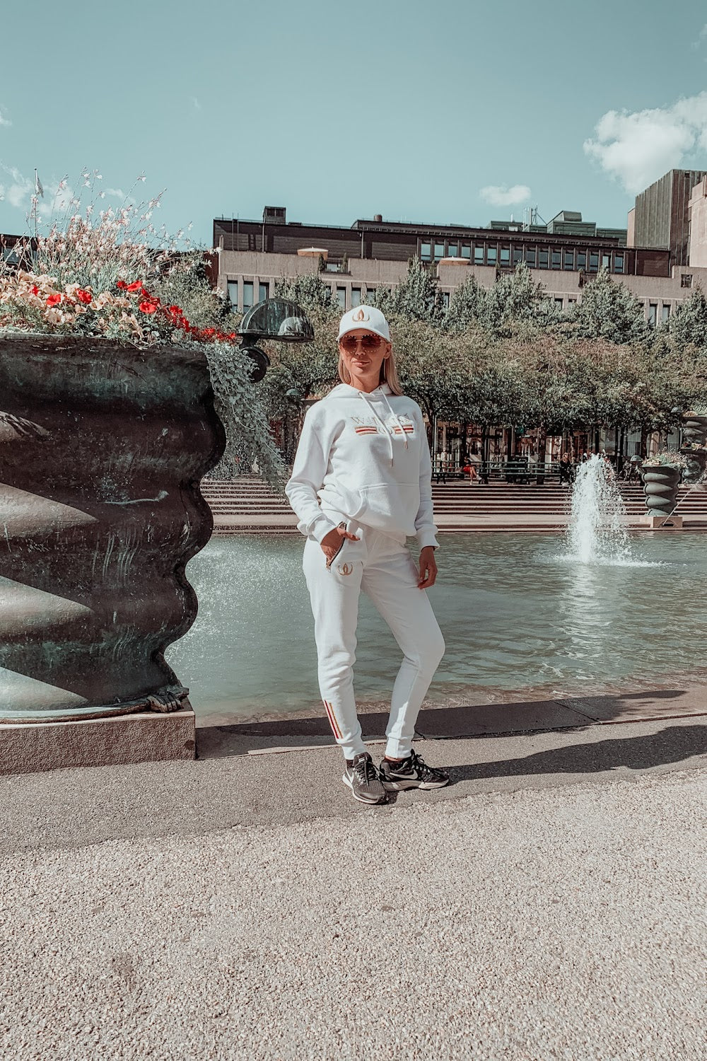 Vor dem Brunnen steht das Model im weißen Hoodie, weißen Joggers und weißer Cap.