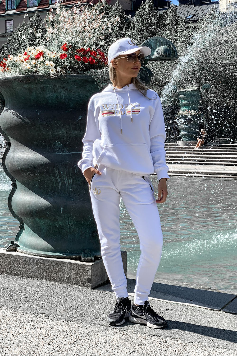 An einer dunklen Urne steht das Model mit den Händen in den Taschen, im weißen Hoodie und weißen Joggers.