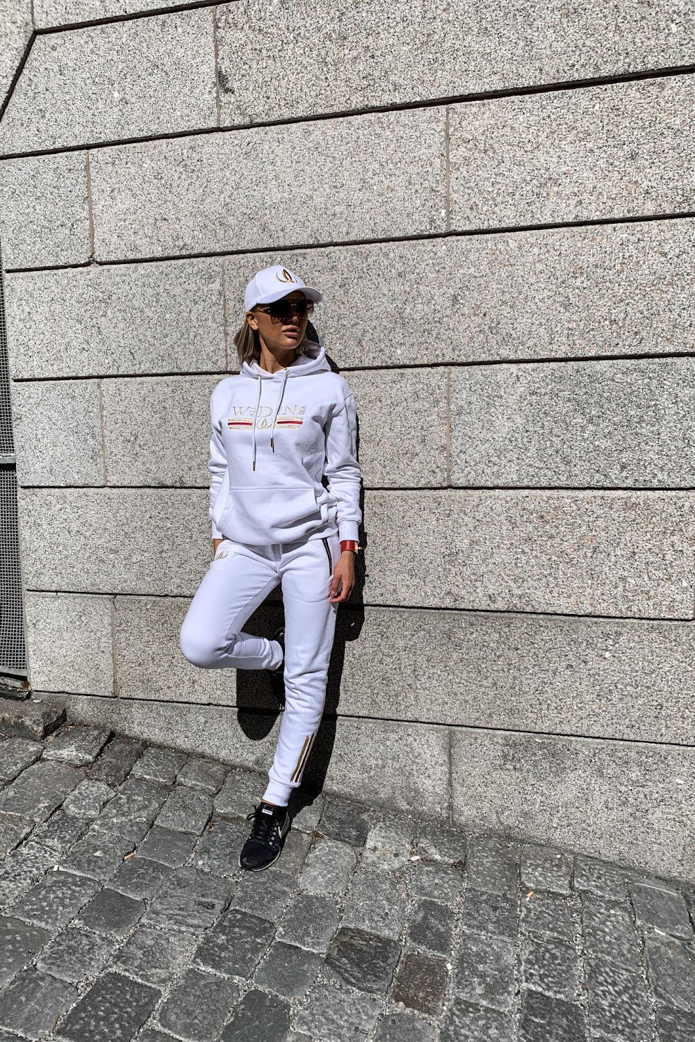 An die Steinmauer lehnt sich das Model mit einem angezogenen Fuß, im weißen Hoodie und weißen Joggers.