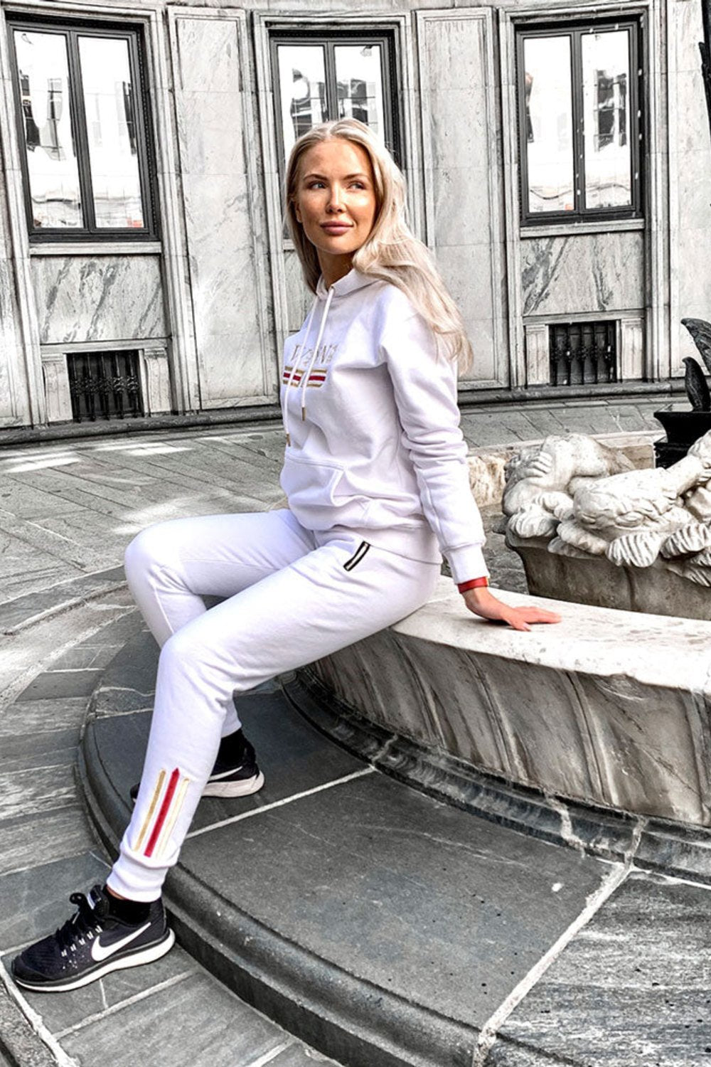 Das Model sitzt an einem Brunnen in der Stadt im weißen Hoodie und weißen Joggern mit Goldstreifen.