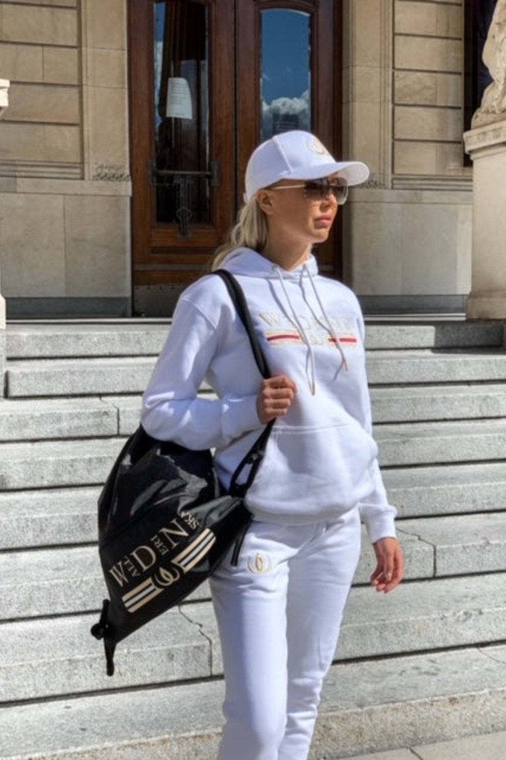 Die Jogger werden zu weißem Hoodie und Cap getragen, mit einem schwarzen Rucksack mit Goldlogo über der Schulter.
