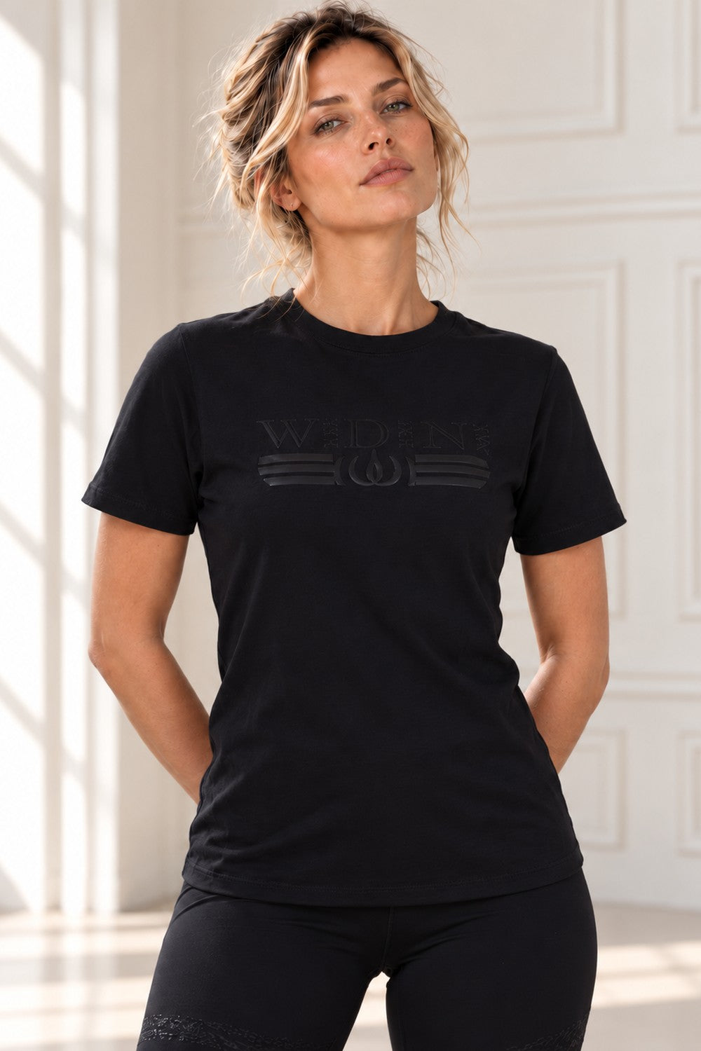 Schwarzes Deluxe Top mit Logo im selben schwarzen Ton, getragen zu schwarzen Leggings.
