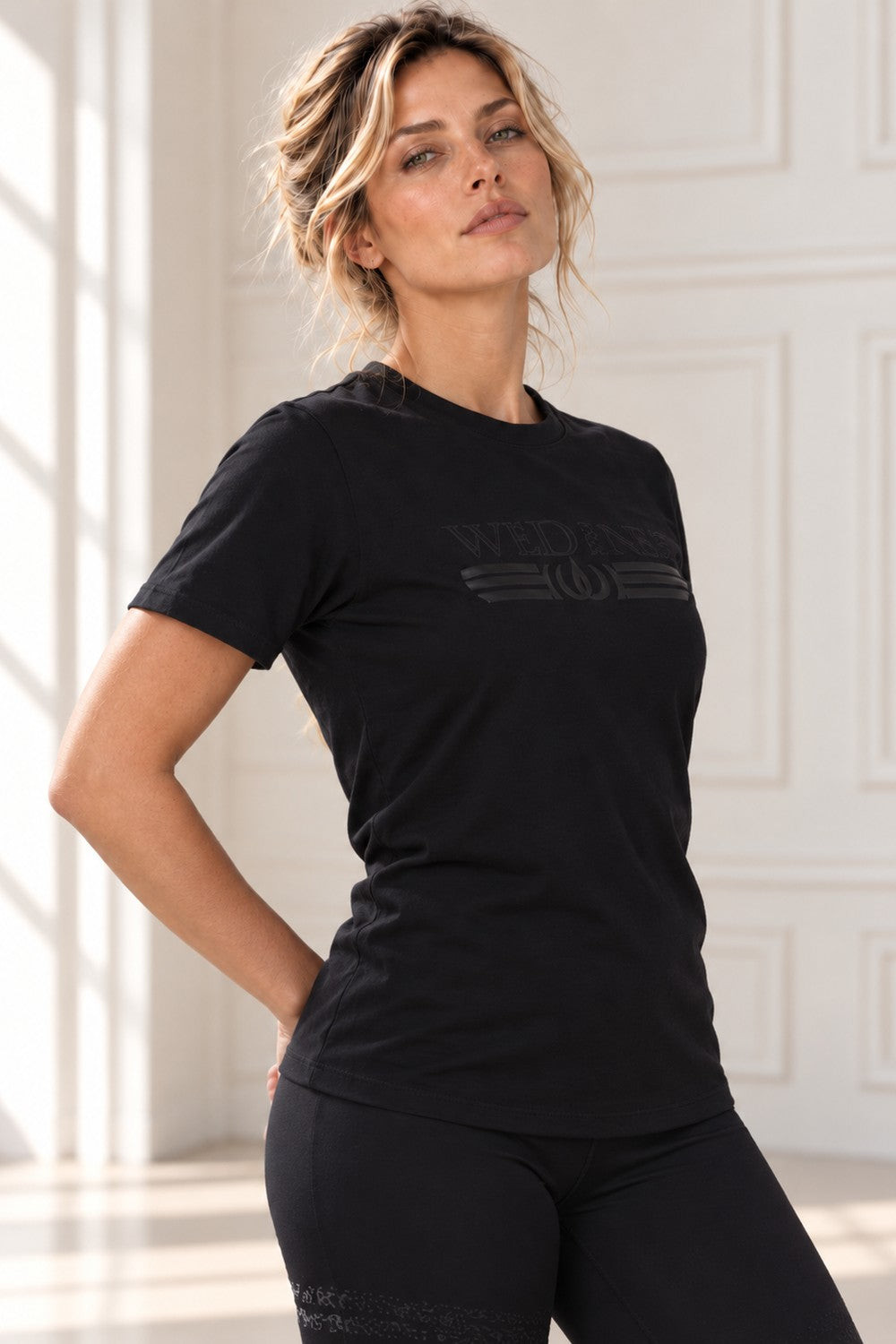 Das Model steht mit der Hand in die Hüfte gestützt im schwarzen Top mit Ton-in-Ton-Logo.
