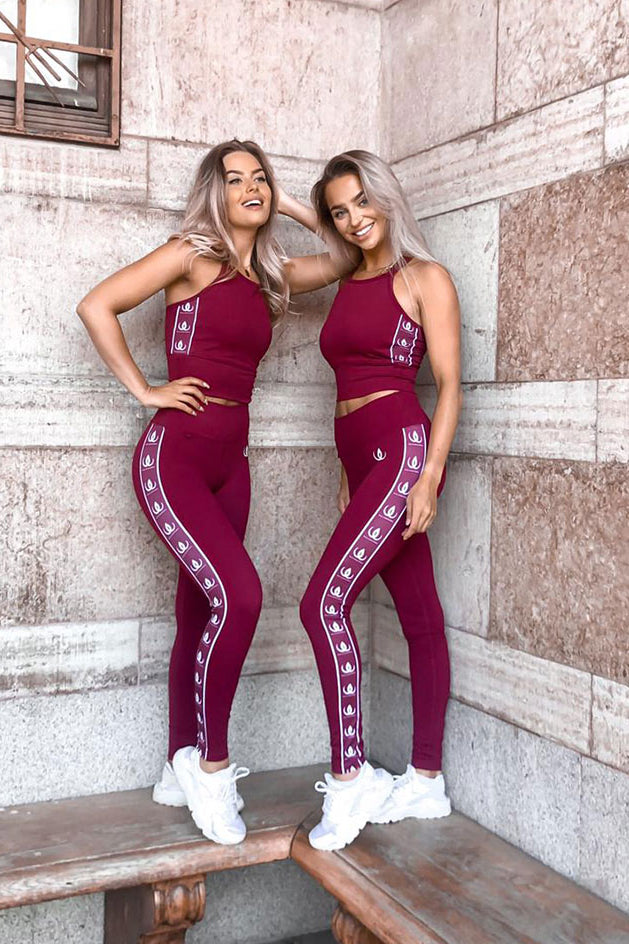 Zwei Models an eine Steinmauer gelehnt in weinroten Croptops und Leggings mit weißen Logobändern.
